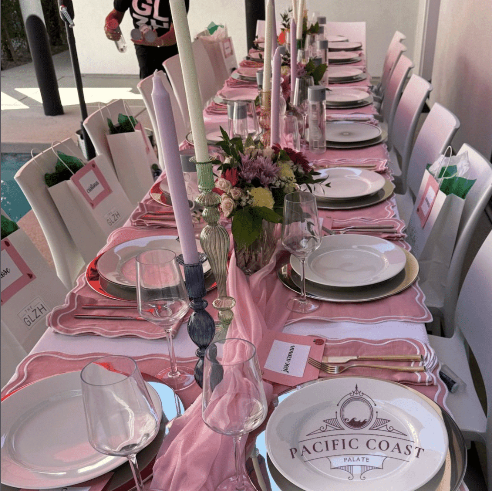 Elegant outdoor dining table set for a dinner party with pink linens, colorful candles, floral centerpieces, personalized place cards, and "Pacific Coast Palate" branded plates.