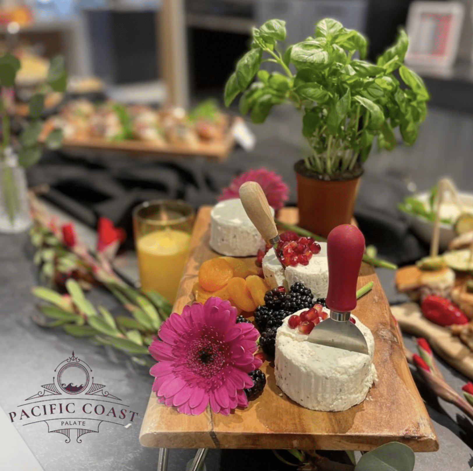 A Pacific Coast Palate cheese board featuring soft white cheeses with pomegranate, blackberries, dried apricots, and a pink daisy. Other catering items are blurred in the background.