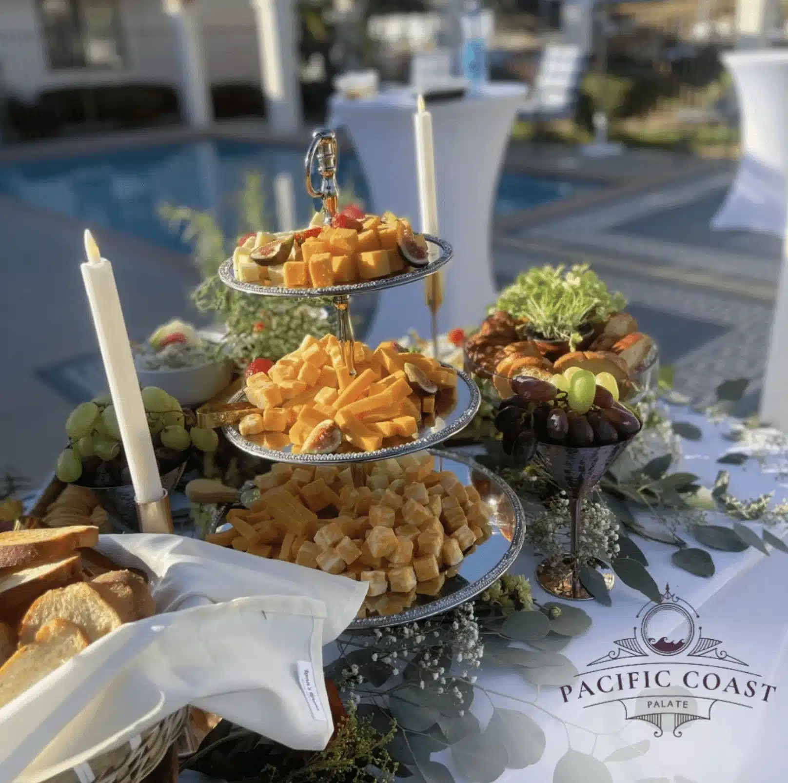 Elegant outdoor catering display by Pacific Coast Palate, featuring a tiered cheese board with figs, strawberries, grapes, and bread by a pool with candles.