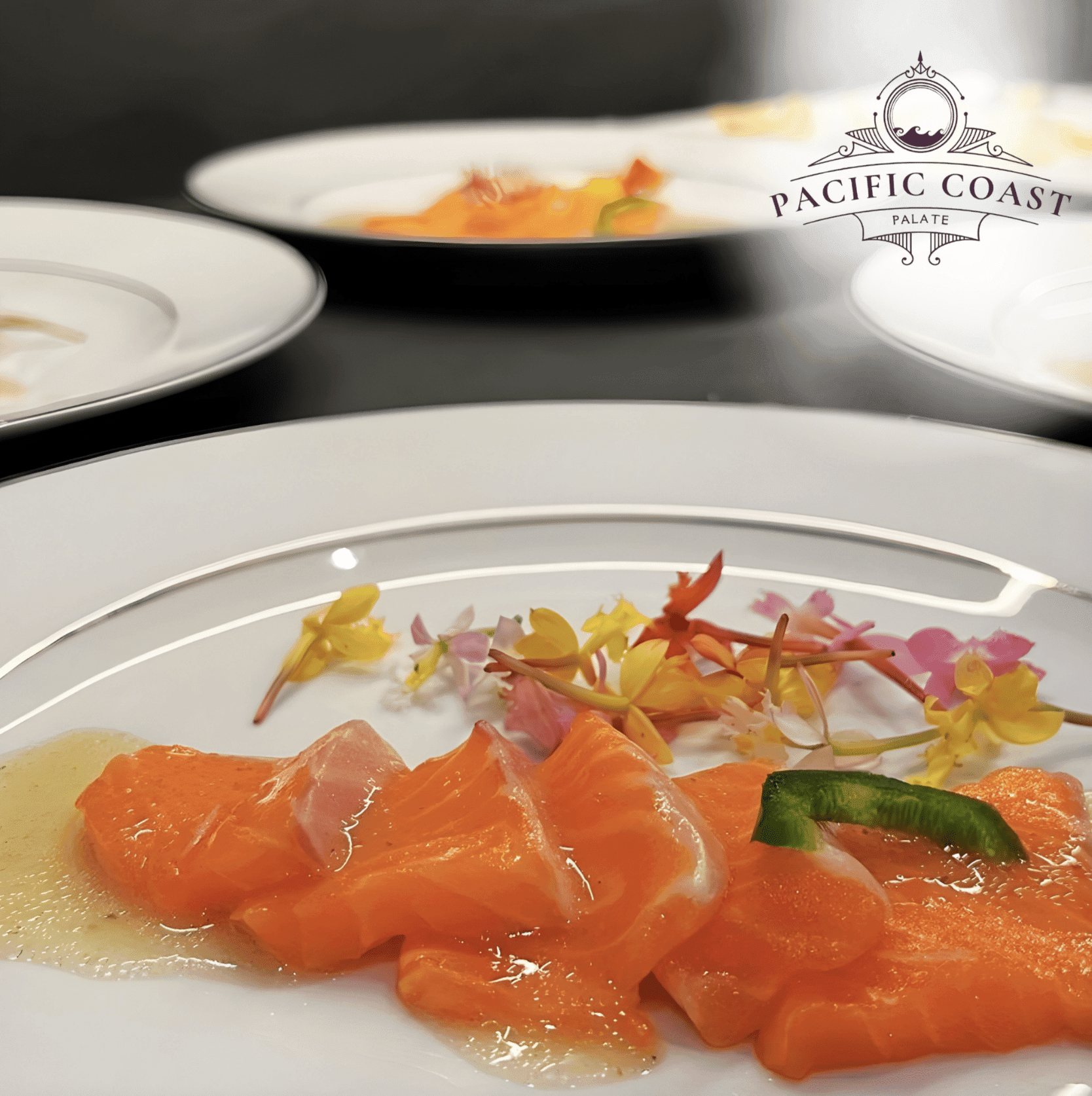 Close-up of elegantly plated salmon sashimi or carpaccio garnished with colorful edible flowers and a green pepper slice, served on a white plate with a silver rim by Pacific Coast Palate.